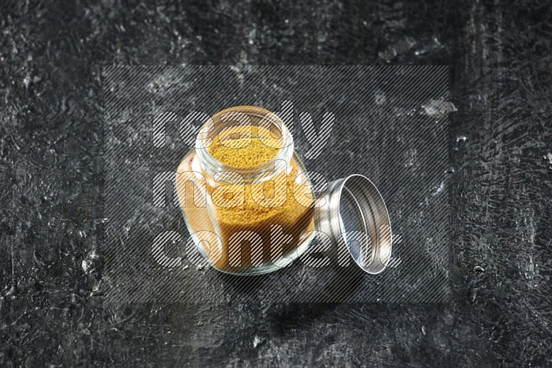 A glass spice jar full of turmeric powder on a textured black flooring