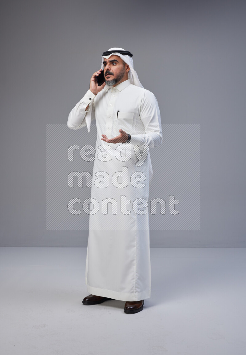 Saudi man Wearing Thob and white Shomag standing talking on phone on Gray background
