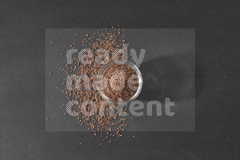 A glass bowl full of flaxseeds surrounded by the seeds on a black flooring