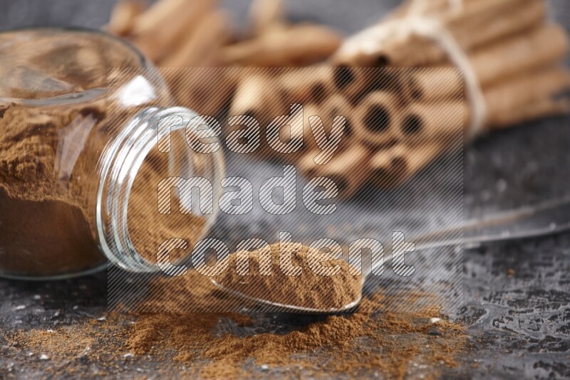 Herbal glass jar full cinnamon powder flipped and a metal spoon full of powder, cinnamon sticks stacked and bounded in the back on textured black background in different angles