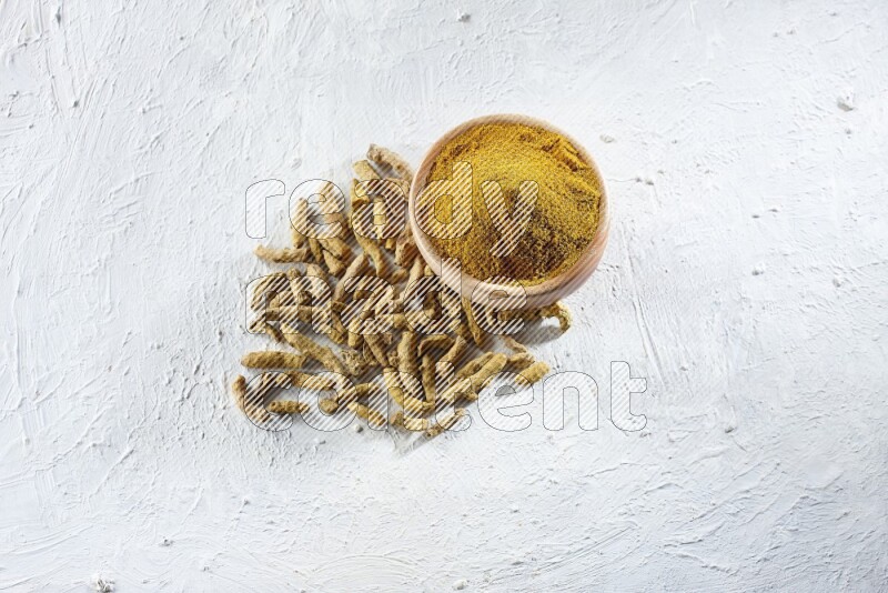 A wooden bowl full of turmeric powder and dried whole fingers beside it on a textured white flooring