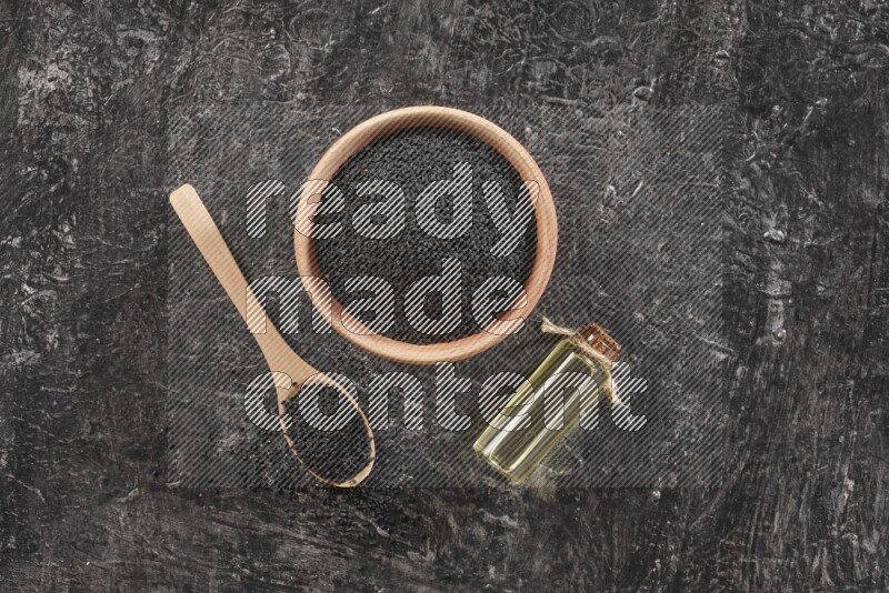 A wooden bowl and spoon full of black seeds and a bottle of black seeds oil on a textured black flooring