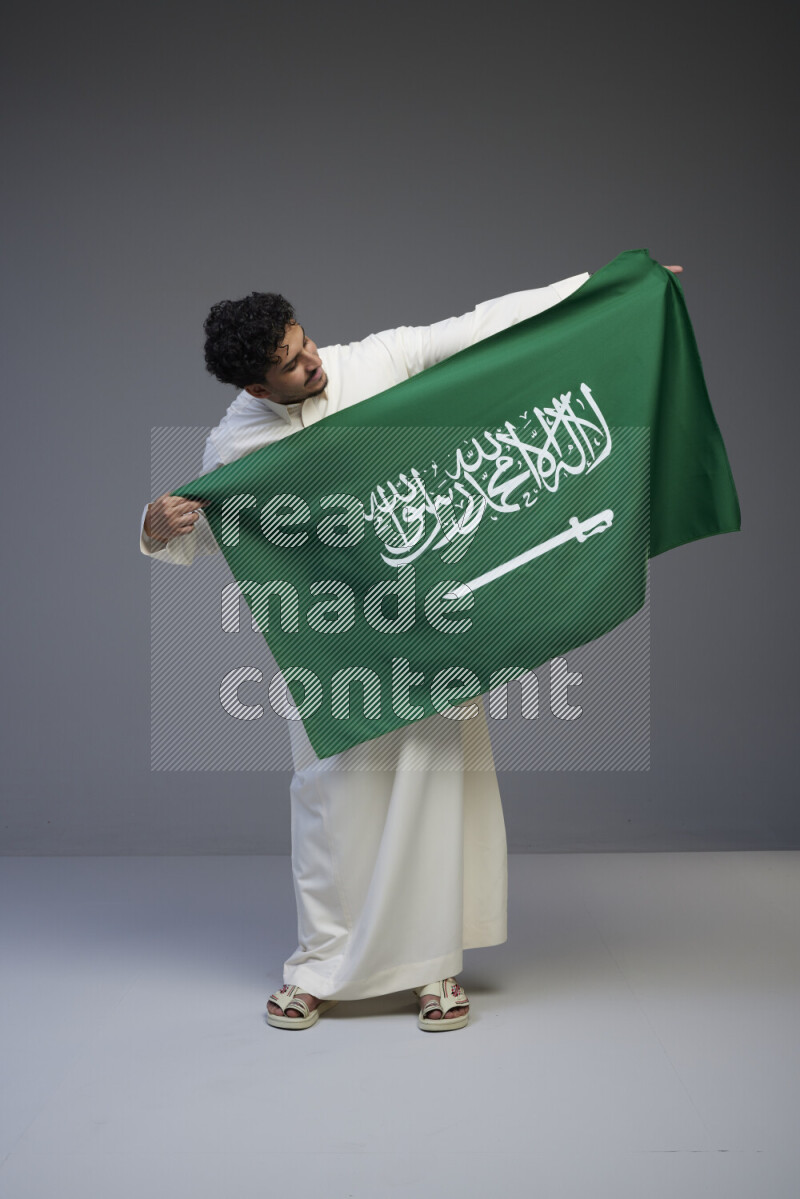 A Saudi man standing wearing thob holding big Saudi flag on gray background