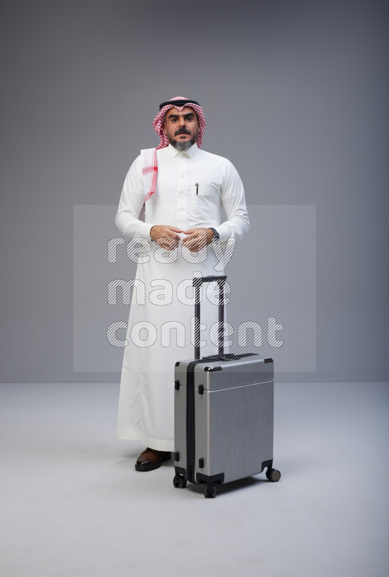 Saudi man wearing Thob and red Shomag standing holding Travel bag on Gray background
