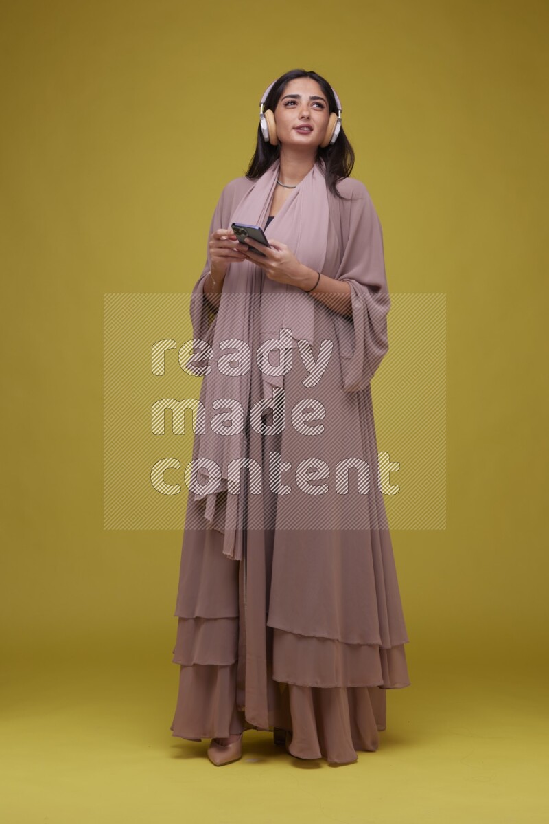 A Saudi woman Listing to Music on a Yellow Background wearing Brown Abaya