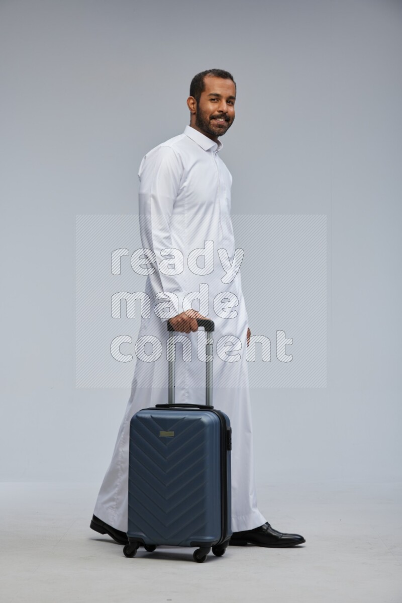Saudi man wearing thob standing holding Travel bag on Gray background