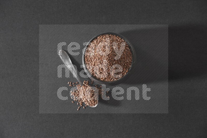 A black pottery bowl full of flaxseeds and a metal spoon full of the seeds on a black flooring