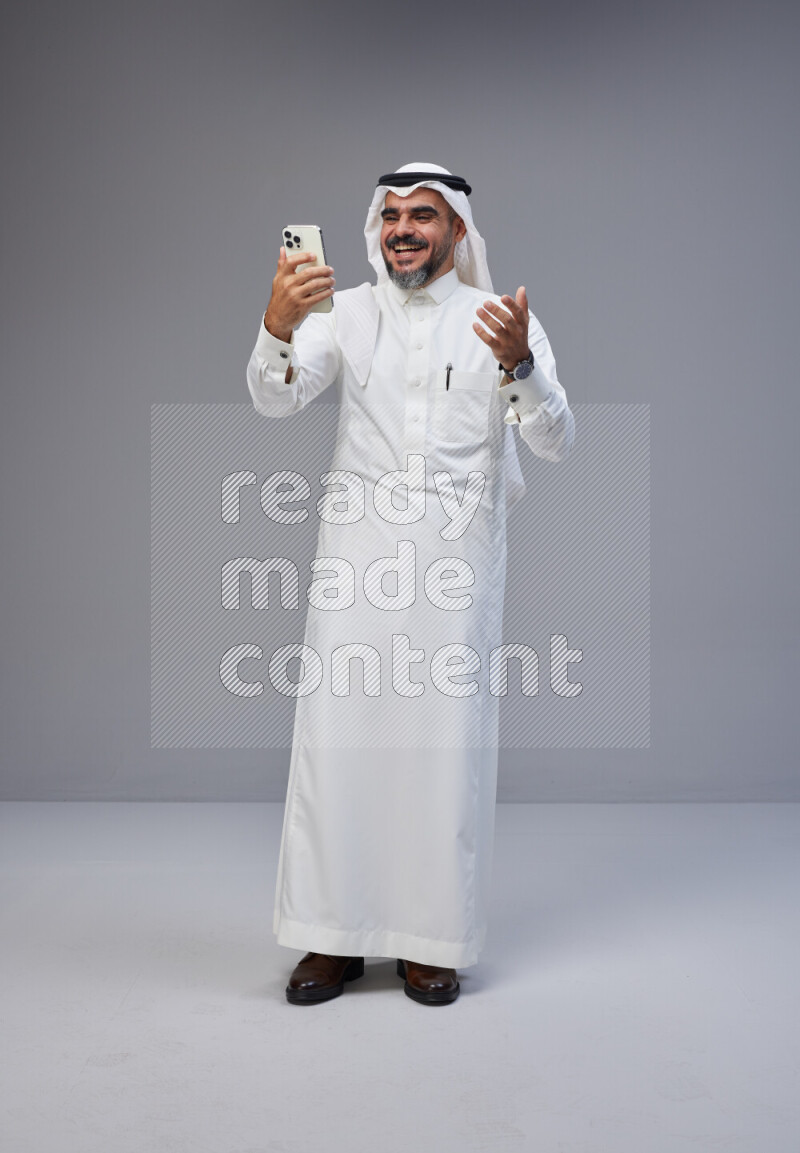 Saudi man Wearing Thob and white Shomag standing texting on phone on Gray background