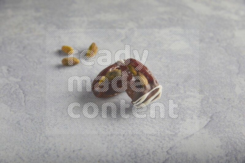 two pistachio stuffed dates covered with dark chocolate with white chocolate drizzle with unroasted pistachios on a light grey background