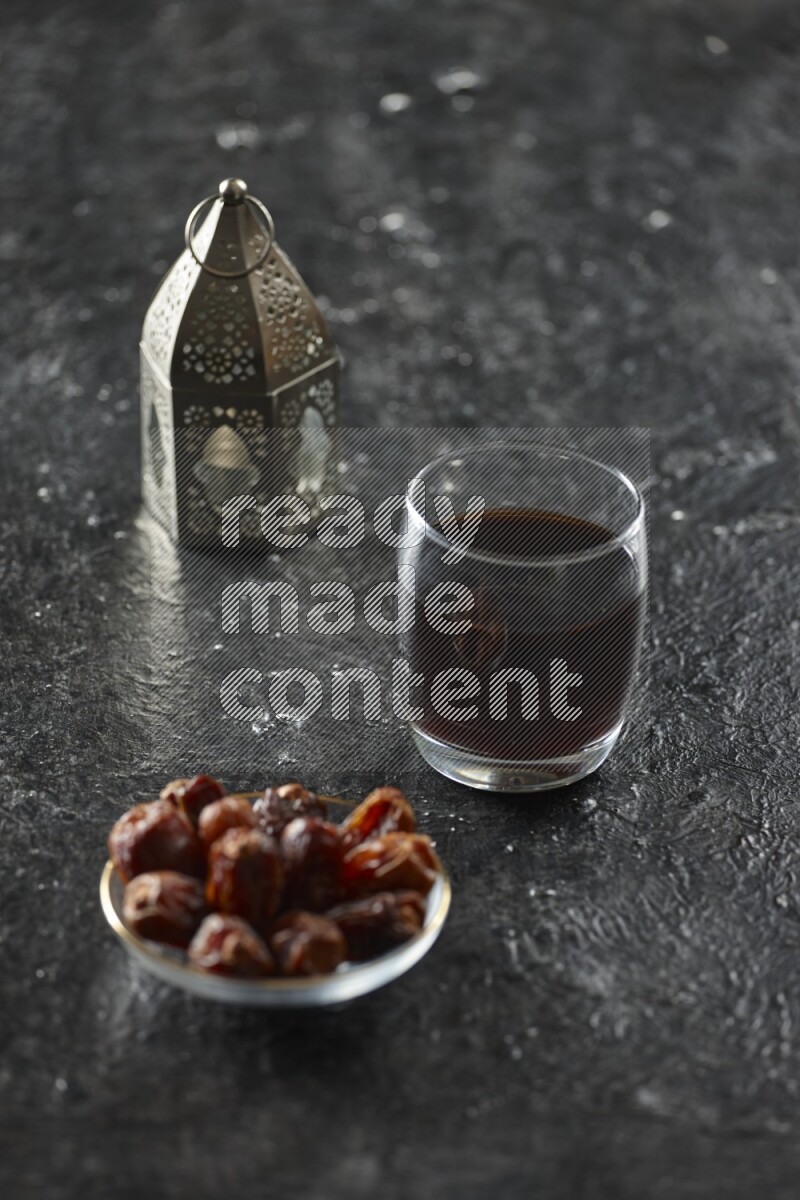 A silver lantern with different drinks, dates, nuts, prayer beads and quran on textured black background