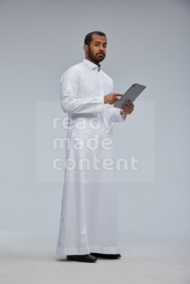 Saudi man wearing Thob standing working on tablet on Gray background