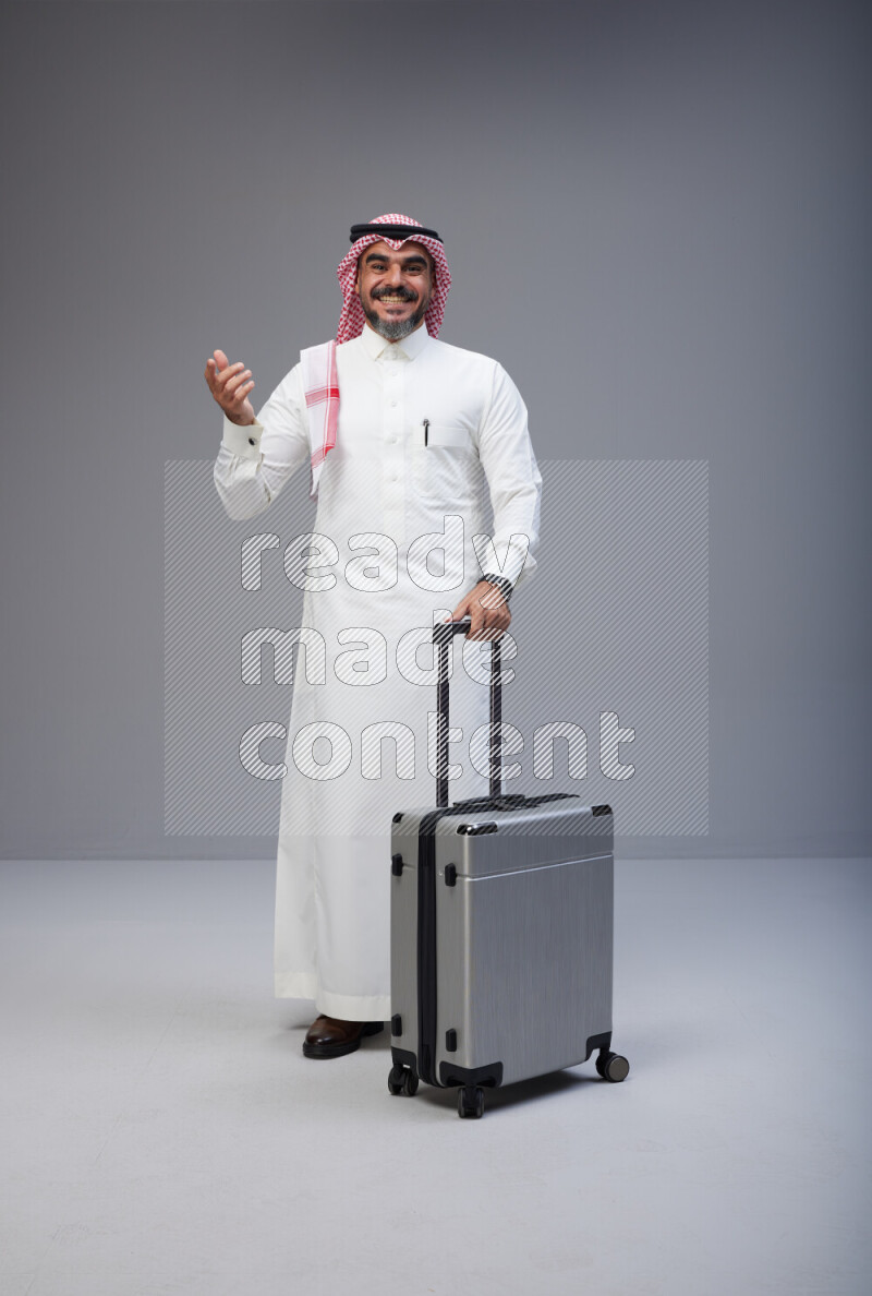 Saudi man wearing Thob and red Shomag standing holding Travel bag on Gray background