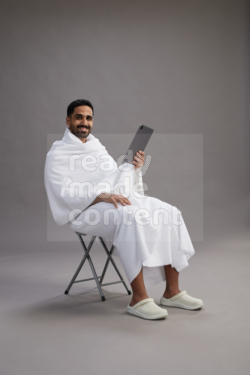 A man wearing Ehram sitting on chair holding a tablet on gray background