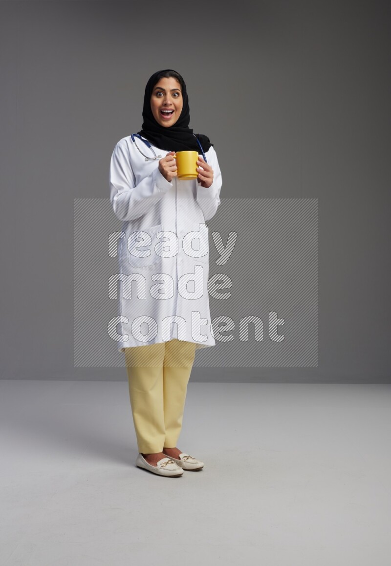 Saudi woman wearing lab coat with stethoscope standing holding mug on Gray background
