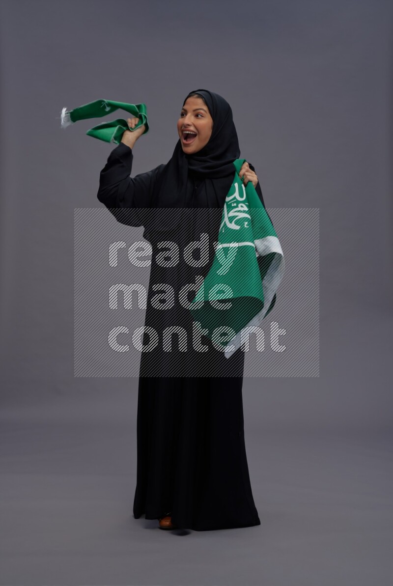 Saudi woman wearing Abaya standing holding Saudi flag on gray background