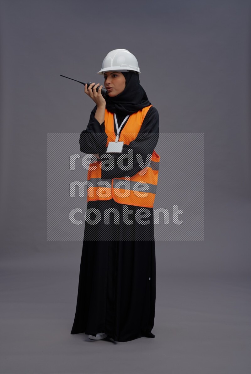 Saudi woman wearing Abaya with engineer vest with neck strap employee badge standing holding walkie-talkie on gray background