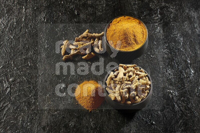 2 pottery black bowls, one full of turmeric powder and the other full of dried turmeric whole fingers on a textured black flooring