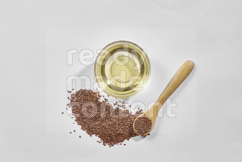 A glass bowl full of flaxseeds oil and wooden spoon full of flaxseeds with spread seeds on a white flooring