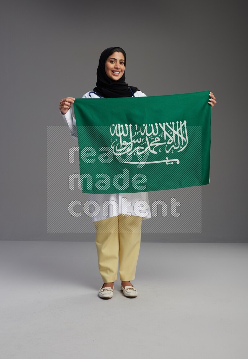 Saudi woman wearing lab coat with stethoscope standing holding Saudi flag on Gray background