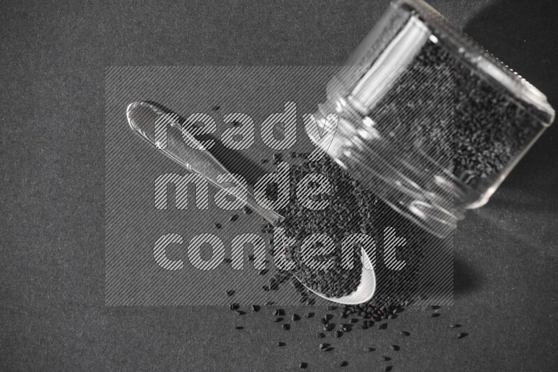 A flipped glass jar full of black seeds and seeds spread out with a metal spoon full of the seeds on a black flooring
