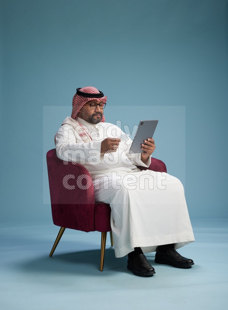 Saudi Man with shimag sitting on chair holding ATM card while working on tablet on blue background
