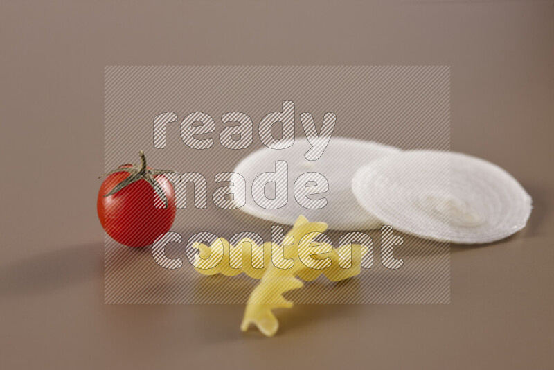 Raw pasta with different ingredients such as cherry tomatoes, garlic, onions, red chilis, black pepper, white pepper, bay laurel leaves, rosemary and cardamom on beige background