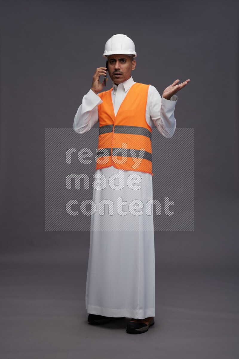 Saudi man wearing thob with engineer vest standing talking on phone on gray background