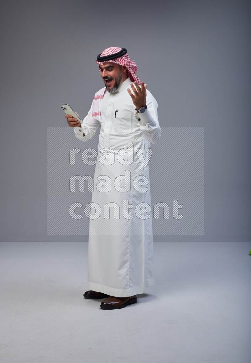 Saudi man Wearing Thob and red Shomag standing texting on phone on Gray background