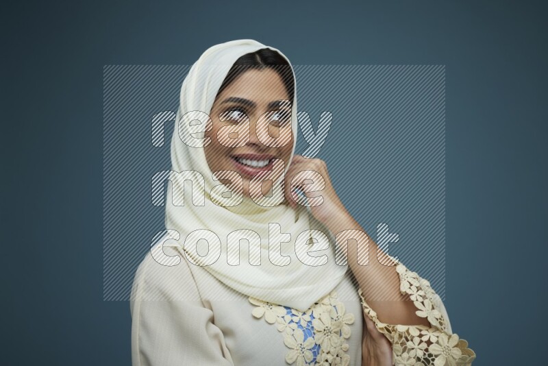 A Saudi woman posing in a blue background wearing an off-white Abaya with hijab