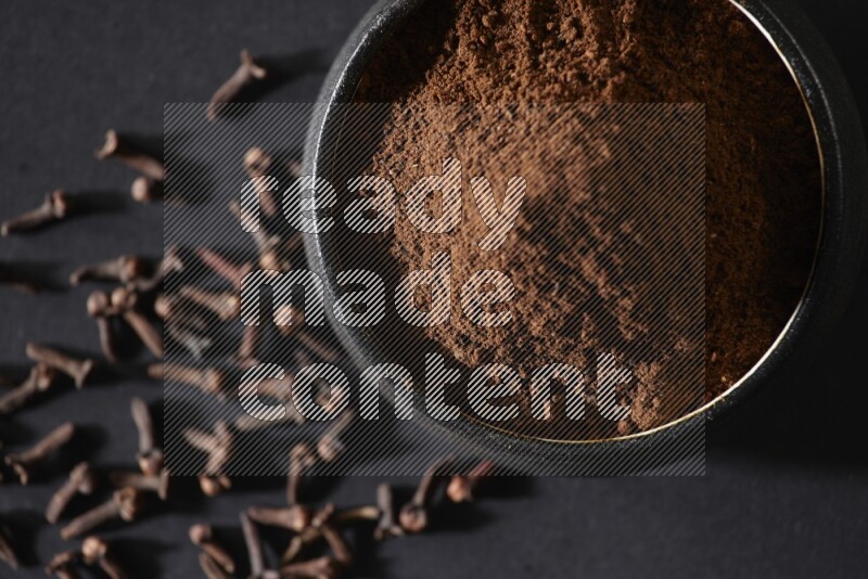 A black pottery bowl full of cloves powder and cloves whole grains on a black flooring