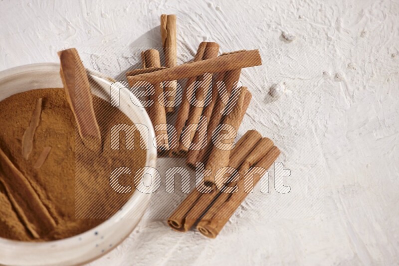 Ceramic bowl full of cinnamon powder with cinnamon sticks on the side on white background