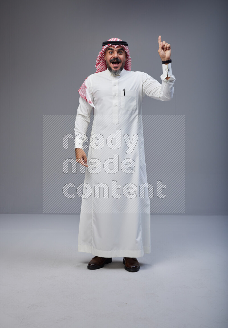 Saudi man Wearing Thob and red white Shomag standing interacting with the camera on Gray background