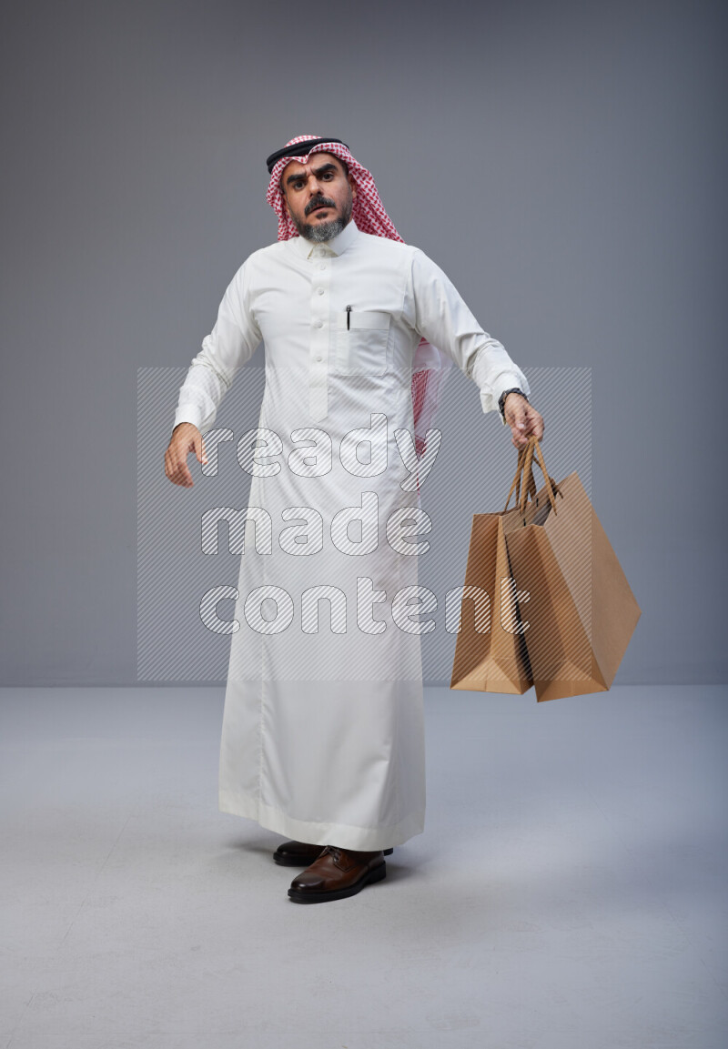 Saudi man Wearing Thob and red Shomag standing holding shopping bag on Gray background