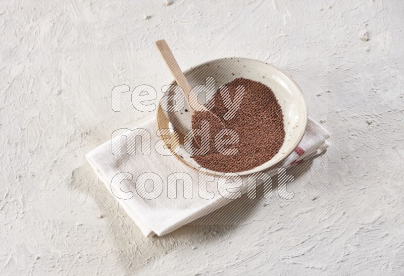 A multicolored pottery plate full of garden cress seeds with a wooden spoon full of the seeds on a napkin on a textured white flooring