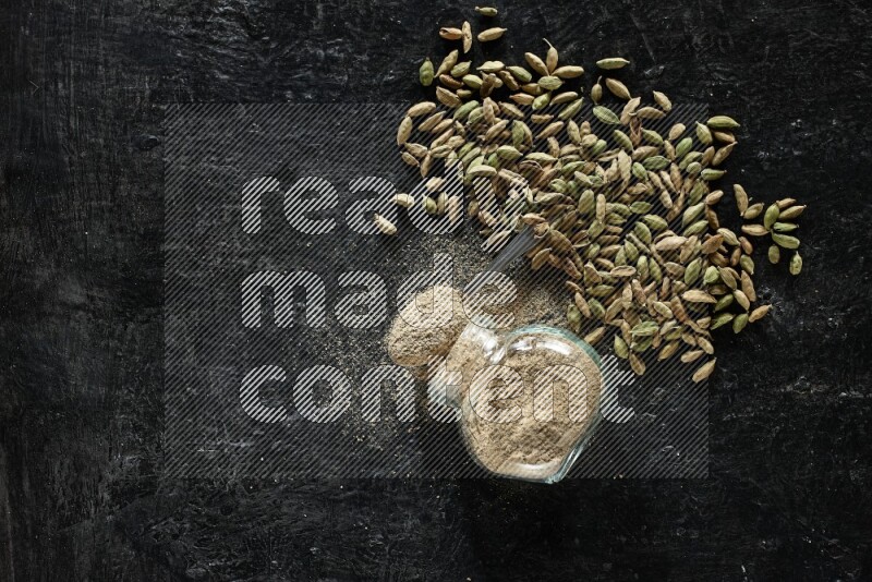 وعاء زجاجي للتوابل مقلوب وملعقة معدنية ممتلئة ببودرة الهيل وحبوب الهيل متناثرة على خلفية سوداء