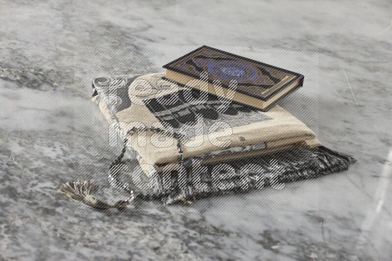 A folded prayer mat with different elements such as quran and prayer beads on grey marble background