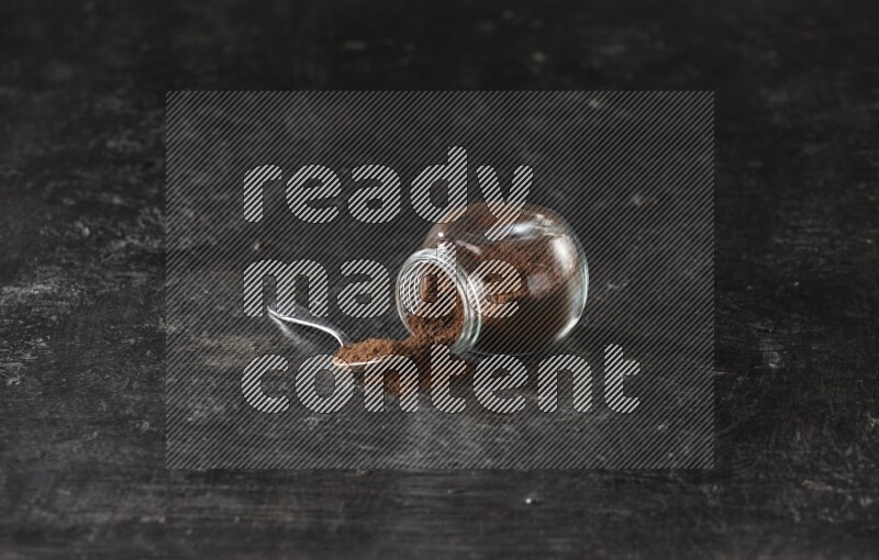 وعاء زجاجي للبهارات مقلوب وملعقة معدنية ممتلئان ببودرة القرنفل على أرضية سوداء