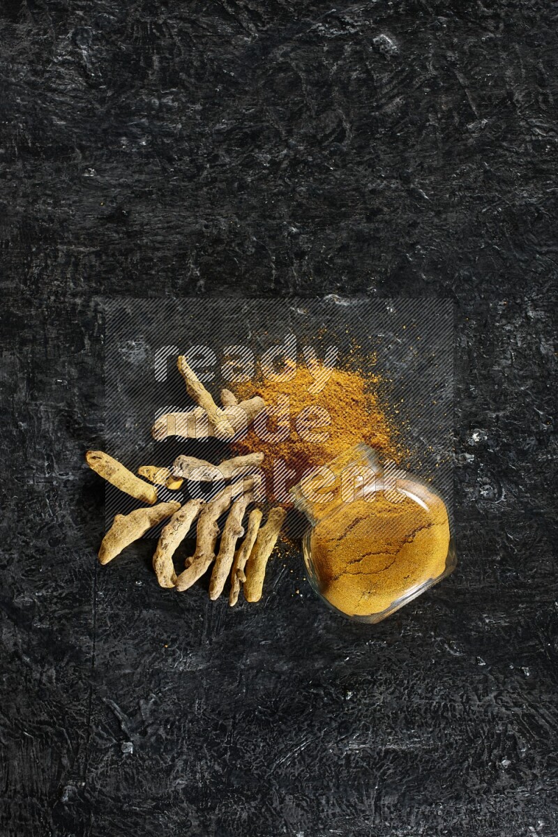 A flipped glass spice jar full of turmeric powder and powder spilled out of it with dried whole fingers on textured black flooring