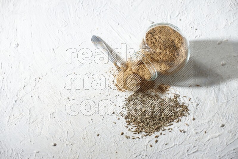 A glass spice jar and metal spoon full of cumin powder and the jar flipped and powder spilled out with cumin seeds on textured white flooring