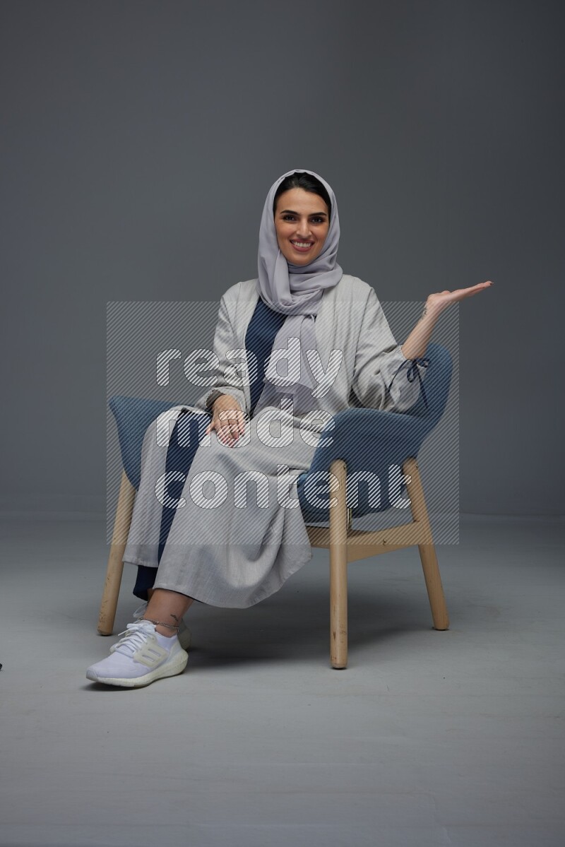 A Saudi woman wearing a light gray Abaya and head scarf sitting on a dark grey chair while making multi hand gestures eye level on a grey background