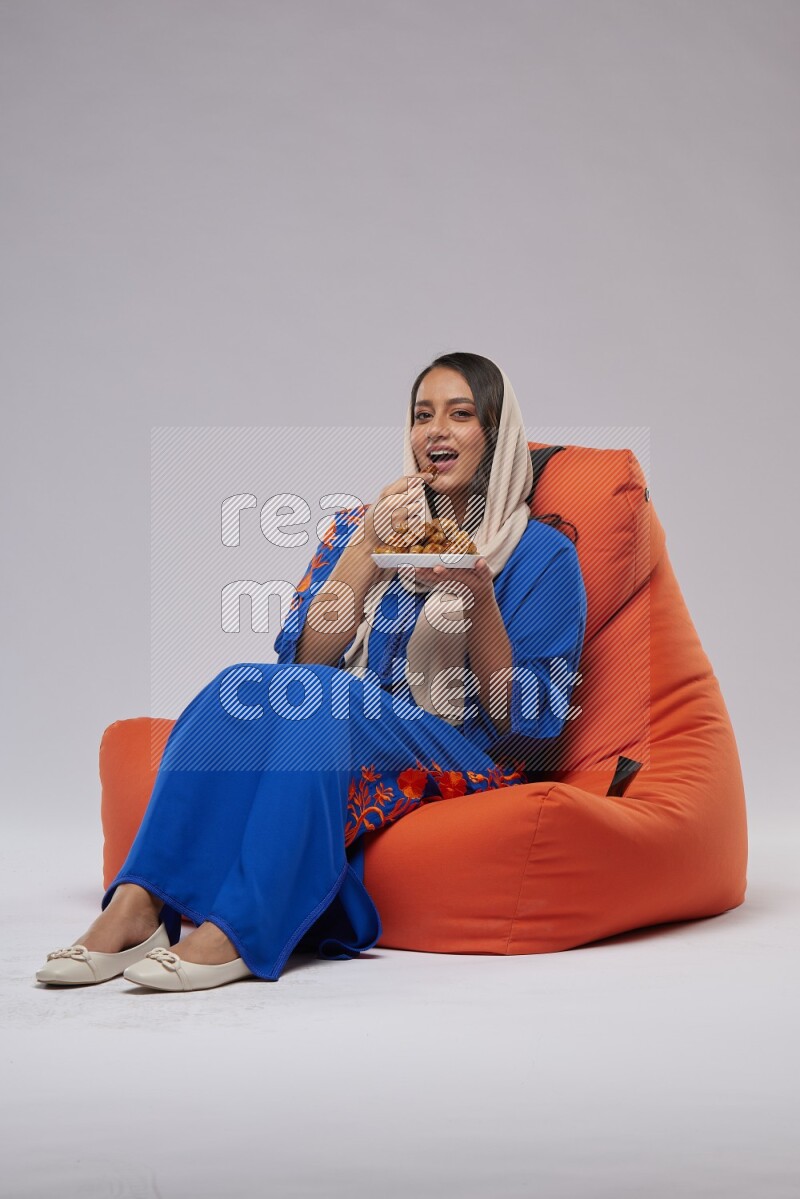 A Woman sitting on an orange beanbag wearing Jalabeya holding a plate of dates