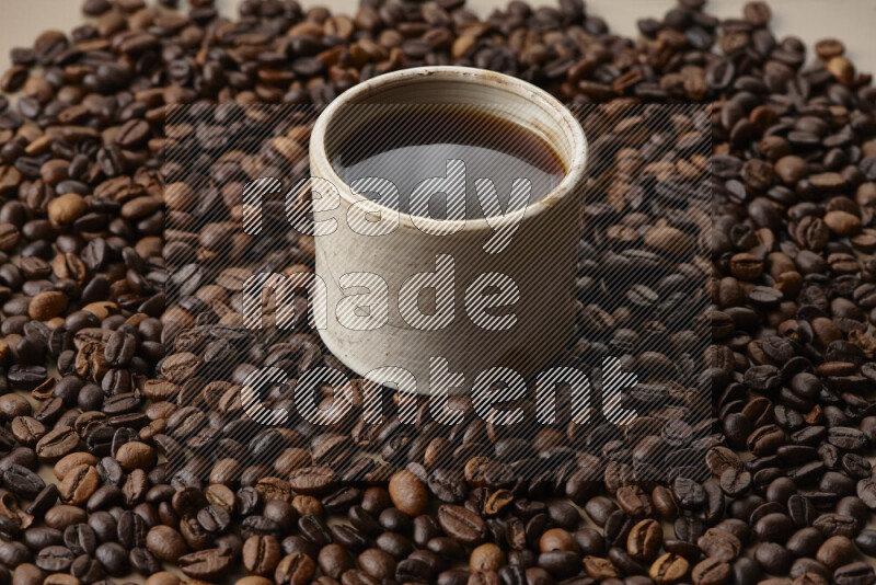 A beige pottery cup of coffee surrounded by roasted coffee beans on beige background