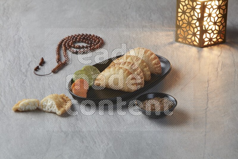 Four fried sambosas in an oval shaped black plate, beside a cut cheese sambosa, a brown misbaha and a golden lantern on a gray background