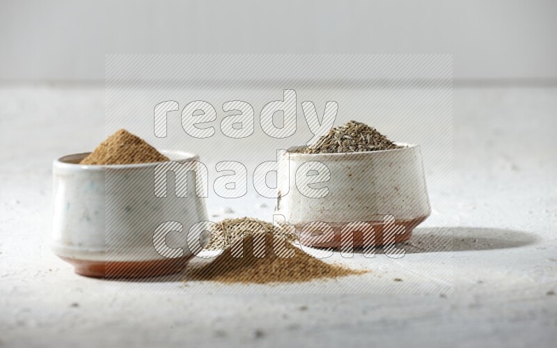2 beige bowls full of cumin seeds and powder with spilled powder and seeds on textured white flooring