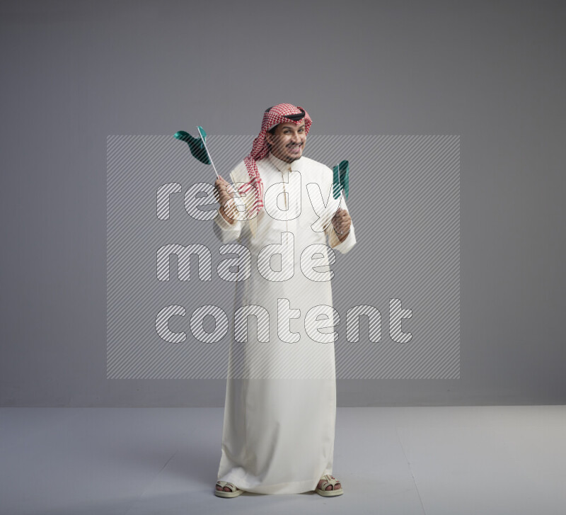 A Saudi man standing wearing thob and red shomag raising small Saudi flag on gray background