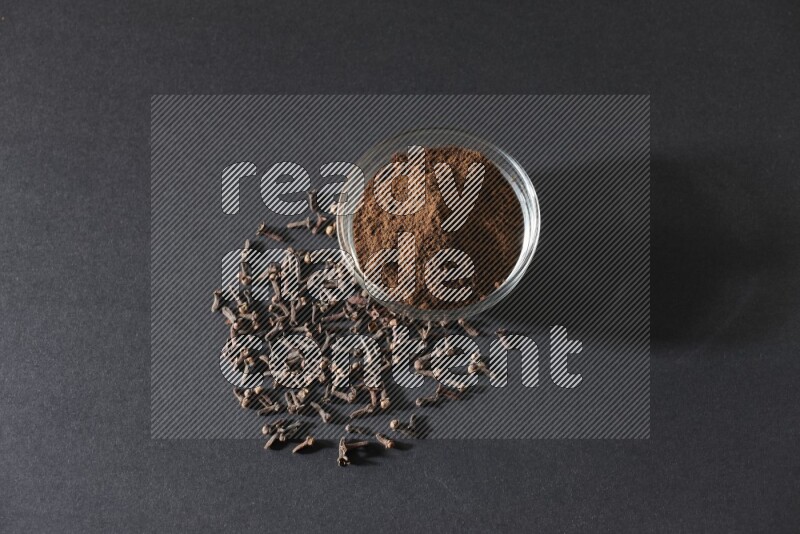 وعاء زجاجي ممتلئ ببودرة القرنفل مع حبوب القرنفل المتناثرة على أرضية سوداء