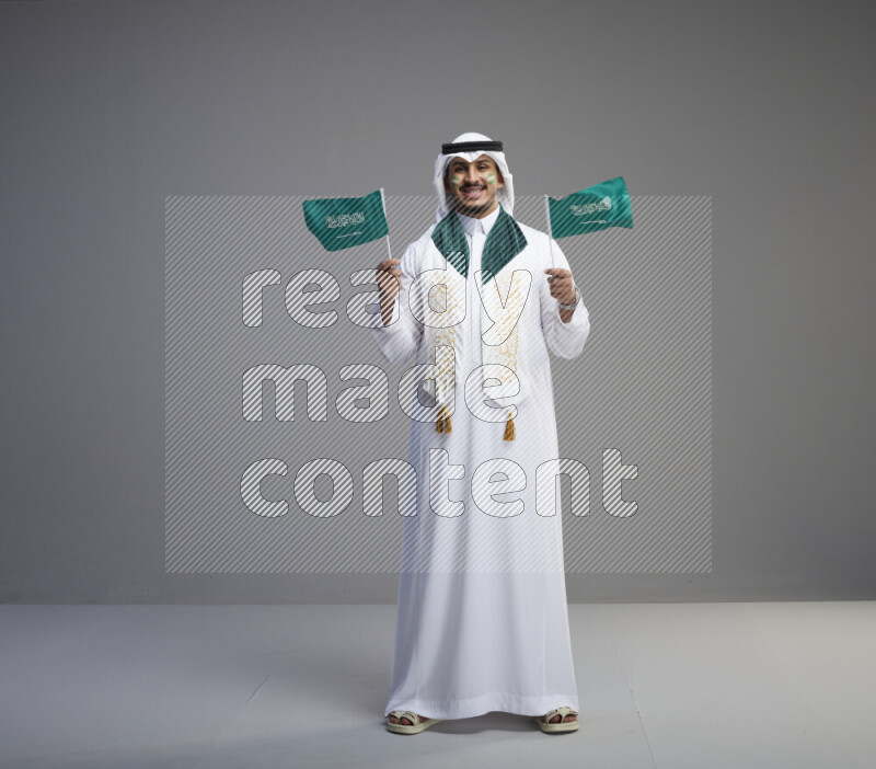 A saudi man standing wearing thob and white shomag with face painting and saudi flag scarf and holding small saudi flag on gray background