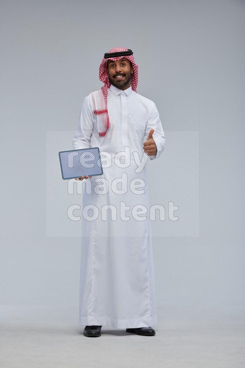 Saudi man Wearing Thob and shomag standing showing tablet to camera on Gray background