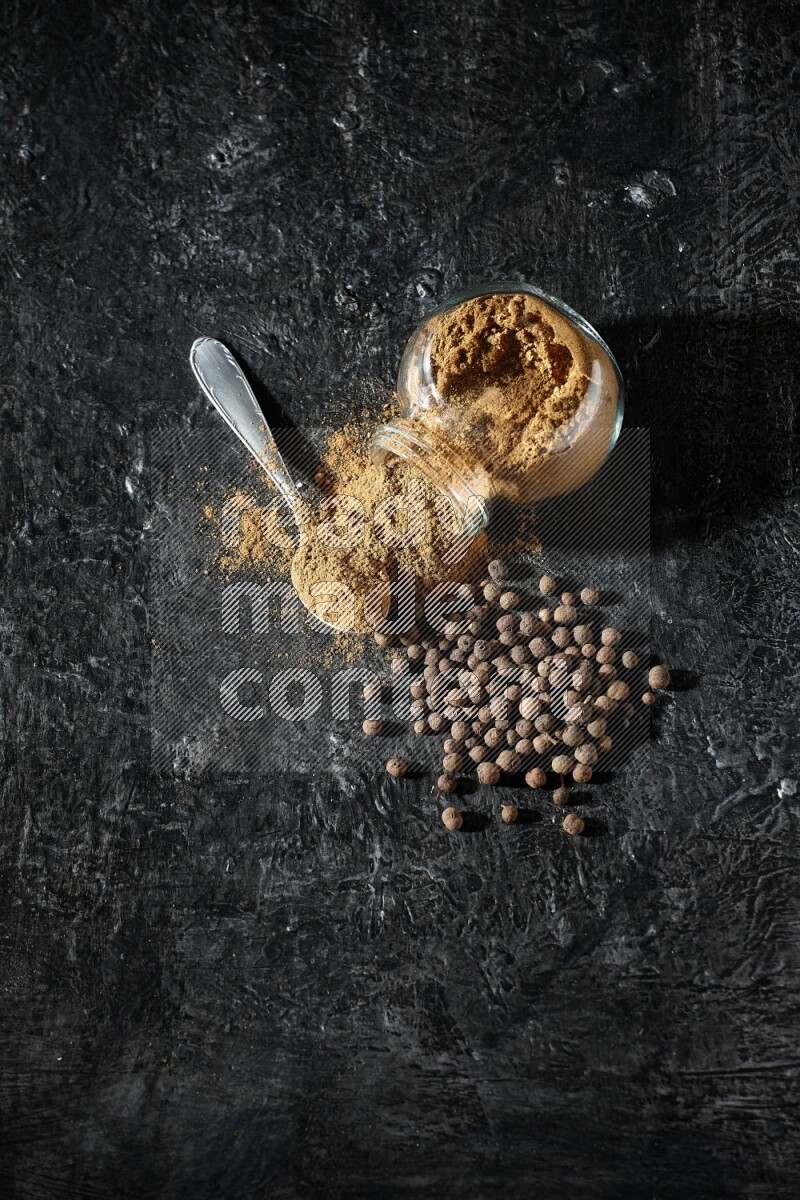 وعاء زجاجي للتوابل مقلوب وملعقة معدنية ممتلئان ببودرة بهار الفلفل الحلو والبودرة تناثرت من الوعاء مع بعض الحبوب على خلفية سوداء