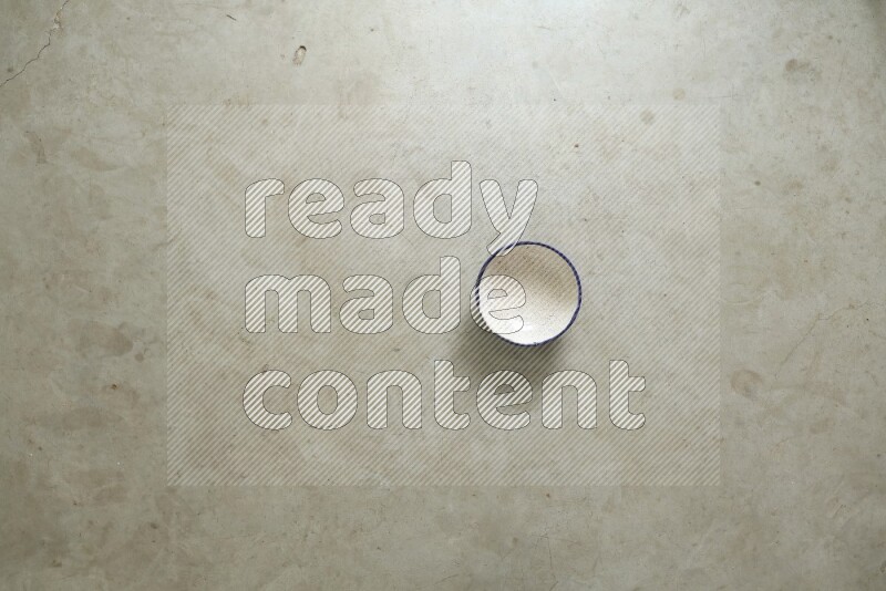 A ceramic bowl beside a carton of eggs and glass bowl full of sugar, vanilla extract, flour on beige marble background